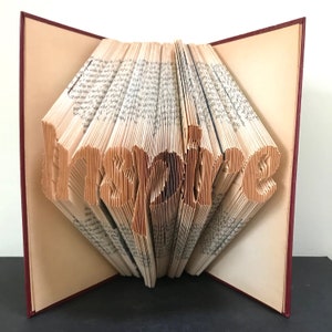 Könnte beinhalten: Ein Buch mit dem Wort "inspire" in die Seiten gefaltet. Das Buch ist geöffnet und die Seiten sind gefaltet, um das Wort zu bilden. Das Buch hat einen roten Einband.