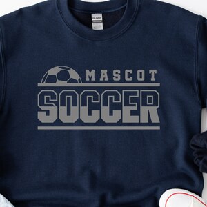 May include: A navy blue sweatshirt with a gray soccer ball graphic and the text "MASCOT SOCCER" in gray.