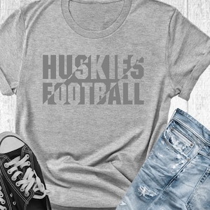 May include: Gray t-shirt with the text "HUSKIES FOOTBALL" printed in a bold, gray font. The text is broken up by diagonal lines.