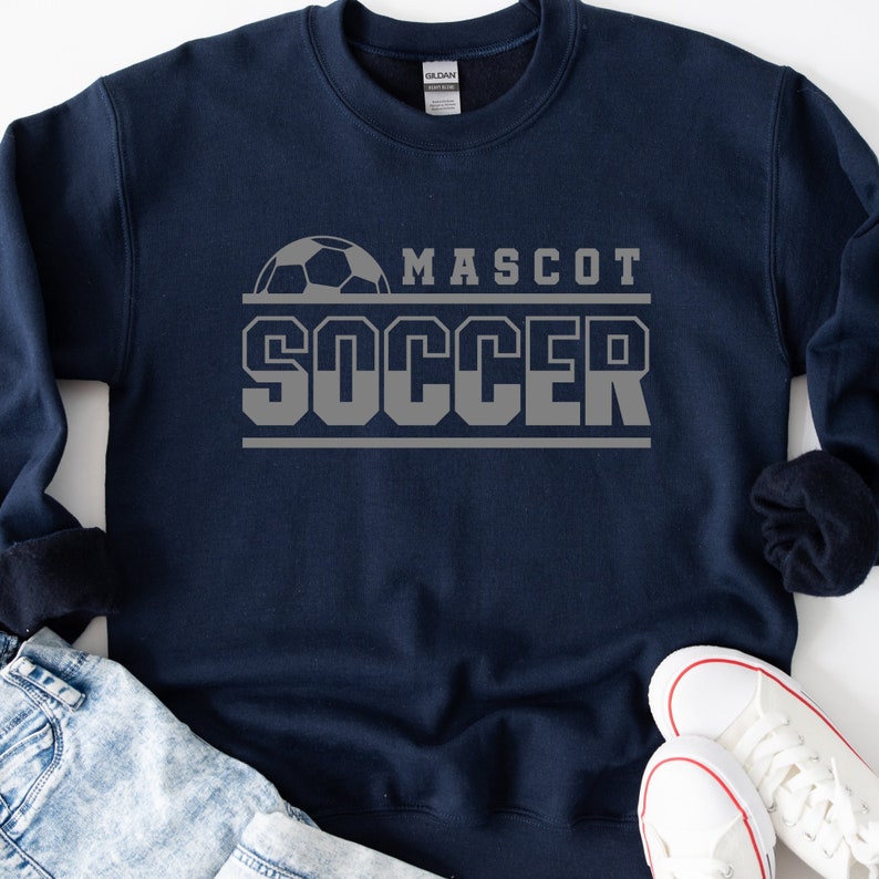 May include: A navy blue sweatshirt with a gray soccer ball graphic and the text "MASCOT SOCCER" in gray.