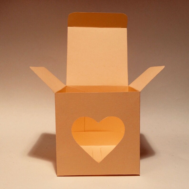 May include: An open, light peach-colored cardboard box with a heart-shaped cutout on the front. The box is designed for packaging small gifts or favors.