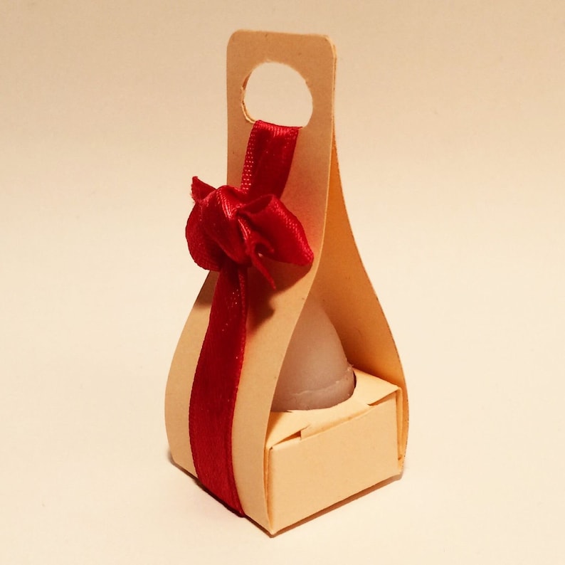 Puede incluir: Una peque&ntilde;a caja de cart&oacute;n de color crema con asa y un lazo de cinta roja. La caja est&aacute; dise&ntilde;ada para contener un objeto peque&ntilde;o y redondo, como una vela o un jab&oacute;n.