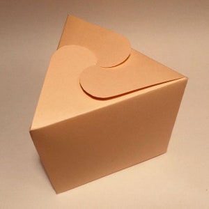 May include: A light peach-colored triangular box with a heart-shaped flap on the top. The box is made of cardboard and has a smooth finish.
