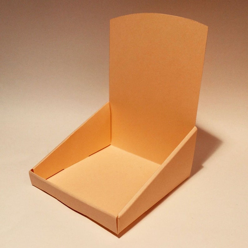 May include: A light peach-coloured cardboard display stand with a back panel and a square base. The stand is designed to hold small items for display.