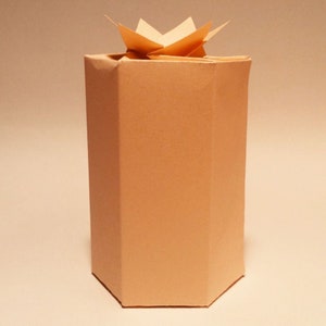 K&ouml;nnte beinhalten: Eine hellbraune Pappbox mit einem gefalteten Papierstern oben. Die Box ist achteckig.