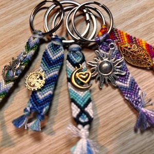 May include: Five colourful friendship bracelet keychains with different charms. The charms include a sun, a crescent moon with a heart, a snowflake, a leaf, and an Om symbol. The keychains are made with colourful embroidery floss and have tassels.