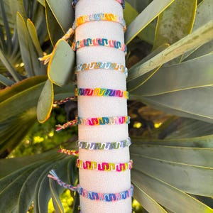 May include: A collection of colorful woven friendship bracelets. Each bracelet features a unique pattern of vibrant threads in shades of blue, pink, yellow, green, and purple. The bracelets are displayed on a white cylindrical stand with a gold top.