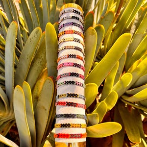 Puede incluir: Una exhibición de coloridas pulseras de la amistad. Las pulseras están apiladas sobre un cilindro blanco con una base dorada, mostrando varios patrones y colores, incluyendo rojo, negro, amarillo y rosa. El fondo presenta hojas de suculentas verdes y amarillas.