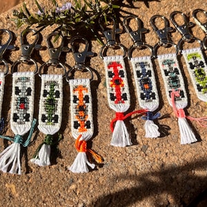 Peut inclure: Plusieurs porte-clés faits à la main avec des motifs de voitures de course brodés aux couleurs vives. Chaque porte-clés est doté d'un fermoir argenté et d'un pompon de couleur assortie. Présentés sur une surface brune texturée.