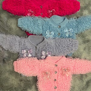 May include: Four handmade crochet cardigans in shades of pink, blue and purple. Each cardigan has a button closure, decorative bows, and a textured, looped design. The cardigans are in hot pink, light blue, lavender, and pastel pink.