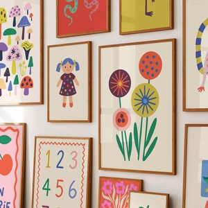 Może przedstawiać: Kolekcja oprawionych wydruk&oacute;w artystycznych o fantazyjnej, kolorowej estetyce. Wydruki przedstawiają r&oacute;żne ilustracje, w tym lalkę, kwiaty, grzyby i liczby. Grafiki są prezentowane w drewnianych ramach.