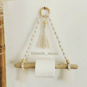 Peut inclure: Un porte-papier toilette en macramé blanc avec une cheville en bois et des perles en bois. Le porte-papier est suspendu à un anneau en bois avec un pompon blanc.