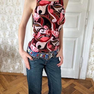 Puede incluir: Una blusa sin mangas con cuello alto y tirantes finos, con un llamativo patrón abstracto de remolinos en tonos rosa, rojo, negro y marrón. La prenda se combina con unos vaqueros de mezclilla azul oscuro y un cinturón azul con bordados florales.