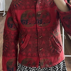 May include: A red jacket with a unique pattern and wooden toggle closures. The jacket features a mandarin collar and two front pockets. The fabric has a textured appearance, and the design includes circular and floral motifs. The jacket is part of a clothing collection.