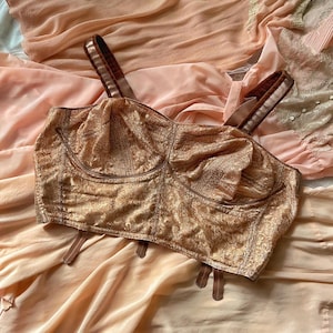 Puede incluir: Un bustier color melocotón de estilo vintage con un diseño texturizado. Cuenta con tirantes y ribetes marrones, sobre un fondo de tela color melocotón. La estética general sugiere lencería o ropa vintage. Una pieza clásica de moda retro.