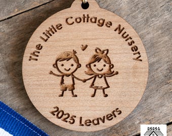 May include: A round wooden ornament with the text "The Little Cottage Nursery 2025 Leavers" and an illustration of two children holding hands. The ornament has a gold-colored string for hanging.