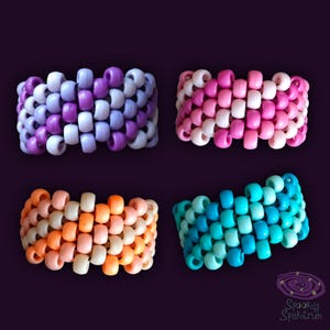 May include: Four colorful beaded bracelets in shades of purple, pink, orange, and blue. Each bracelet is made of small, round beads woven together. The bracelets are displayed on a dark purple background.