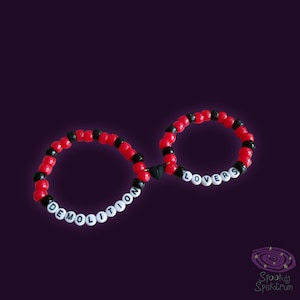 May include: Two beaded bracelets in red, black, and white. One bracelet spells "DEMOLITION" and the other spells "LOVERS". A black heart connects the two bracelets. The background is a dark purple.