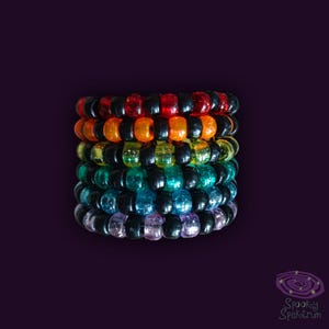 Könnte beinhalten: Ein Stapel bunter Perlenarmbänder in Regenbogenfarben, darunter Rot, Orange, Gelb, Grün, Blau und Lila. Jedes Armband hat eine Mischung aus transparenten und schwarzen Perlen. Auf dunklem lila Hintergrund.