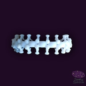 Può includere: Un braccialetto bianco articolato con un motivo ripetuto di forme arrotondate. Il braccialetto è realizzato in un materiale di colore chiaro ed è posto su uno sfondo viola scuro. Il design ricorda una spina dorsale o una serie di elementi collegati.