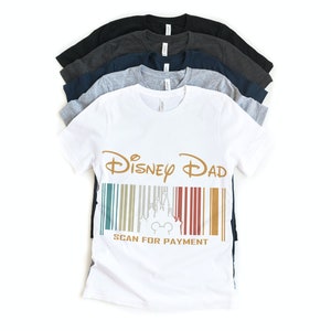 May include: White t-shirt with a gold graphic that says "Disney Dad" and a barcode with a silhouette of Cinderella's castle and Mickey Mouse. The text below the barcode says "Scan for Payment".