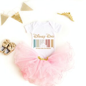 May include: White baby bodysuit with the text "Disney Dad" and a barcode featuring a Mickey Mouse head. The bodysuit is paired with a pink tulle tutu and gold baby shoes.