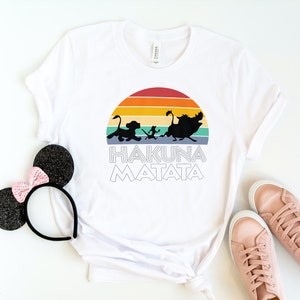 May include: White t-shirt with a retro sunset graphic featuring the Lion King characters Simba, Timon, and Pumbaa. The text "Hakuna Matata" is printed below the graphic.