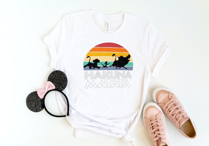 May include: White t-shirt with a retro sunset graphic featuring the Lion King characters Simba, Timon, and Pumbaa. The text "Hakuna Matata" is printed below the graphic.