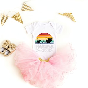 May include: White baby onesie with a rainbow sunset graphic and the words "Hakuna Matata" printed on it. The onesie is paired with a pink tulle tutu and gold baby shoes.