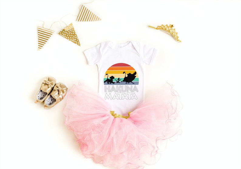 May include: White baby onesie with a rainbow sunset graphic and the words "Hakuna Matata" printed on it. The onesie is paired with a pink tulle tutu and gold baby shoes.