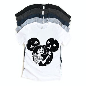 May include: A white t-shirt with a black silhouette of a woman with curly hair inside a Mickey Mouse head shape. The woman is wearing a floral patterned dress and has butterflies around her.