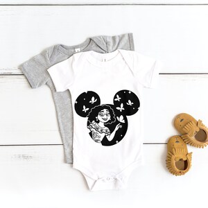 May include: A white baby onesie with a black and white illustration of a woman with curly hair inside a Mickey Mouse silhouette. The woman is holding a butterfly and there are butterflies flying around her. The onesie is on a white wooden surface.