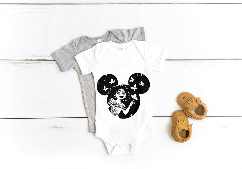 May include: A white baby onesie with a black and white illustration of a woman with curly hair inside a Mickey Mouse silhouette. The woman is holding a butterfly and there are butterflies flying around her. The onesie is on a white wooden surface.