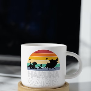 May include: White ceramic mug with a colorful retro sunset design featuring the characters Simba, Timon, and Pumbaa from Disney's The Lion King. The mug has the text "Hakuna Matata" printed on the front.