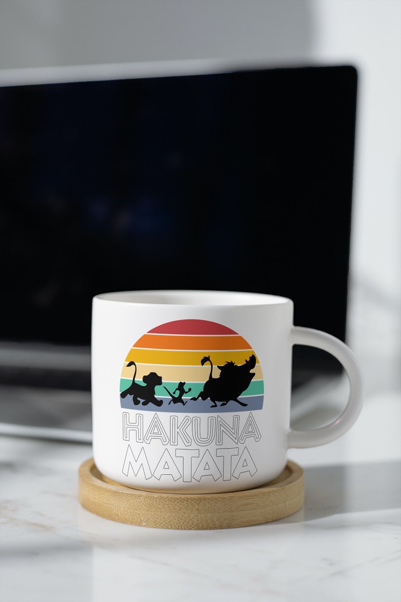 May include: White ceramic mug with a colorful retro sunset design featuring the characters Simba, Timon, and Pumbaa from Disney's The Lion King. The mug has the text "Hakuna Matata" printed on the front.