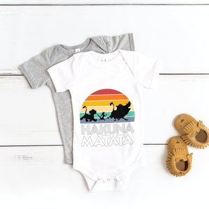 May include: White baby onesie with a rainbow sunset graphic and the text "Hakuna Matata" in black. The onesie is on top of a gray onesie. There are two brown moccasins on a white wooden surface.