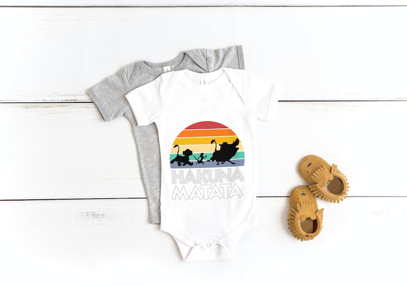 May include: White baby onesie with a rainbow sunset graphic and the text "Hakuna Matata" in black. The onesie is on top of a gray onesie. There are two brown moccasins on a white wooden surface.