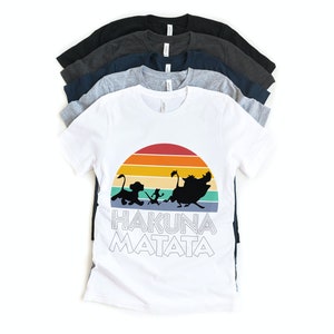 May include: A white t-shirt with a retro sunset graphic and the words "Hakuna Matata" printed on it. The graphic features silhouettes of Simba, Timon, and Pumbaa from the Disney movie The Lion King.