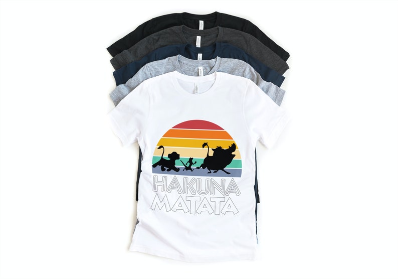 May include: A white t-shirt with a retro sunset graphic and the words "Hakuna Matata" printed on it. The graphic features silhouettes of Simba, Timon, and Pumbaa from the Disney movie The Lion King.