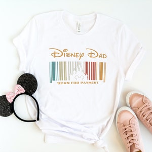 May include: White t-shirt with a gold graphic that says "Disney Dad" and a barcode with a silhouette of Cinderella's castle and Mickey Mouse. The text below the barcode says "Scan for Payment".