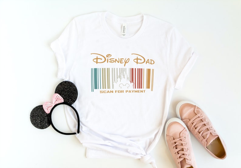 May include: White t-shirt with a gold graphic that says "Disney Dad" and a barcode with a silhouette of Cinderella's castle and Mickey Mouse. The text below the barcode says "Scan for Payment".