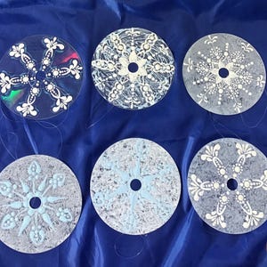 May include: Six decorative CDs with snowflake designs. The snowflakes are painted in white and light blue on a variety of backgrounds, including clear and gray. The CDs are arranged on a blue surface.