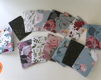 May include: A collection of 12 small notebooks with floral and botanical designs. The notebooks are arranged in a semi-circle on a white surface. The covers feature a variety of floral patterns, including roses, succulents, and leaves.