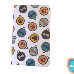 May include: A white notebook with a repeating pattern of dog tags. The tags feature paw prints and bones in various colors, including yellow, red, and blue. The notebook has a rounded corner design.
