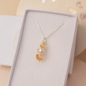 May include: A silver necklace with a pendant of yellow gemstones and a pearl, wrapped in silver wire. The necklace is displayed in a white box, set against a neutral background.