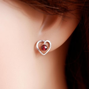 May include: A silver heart-shaped stud earring with a small, round, red gemstone set in the centre. The earring is worn on a person's ear, with the focus on the jewellery.