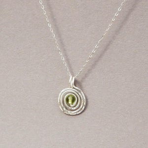 May include: A silver pendant necklace featuring a spiral design. The pendant has a textured, hammered appearance and a small, round, green gemstone at its center. The chain is delicate and silver, and the background is a solid gray.
