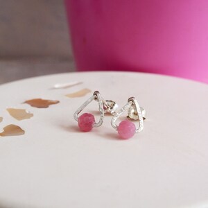 May include: A pair of silver earrings with pink gemstone accents. The earrings feature a triangular design with a small pink gemstone in the centre of each triangle.