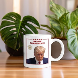 May include: White ceramic mug with a red and white graphic of a man with blonde hair and a serious expression. The graphic includes the text "NEVER SURRENDER!" and five stars.