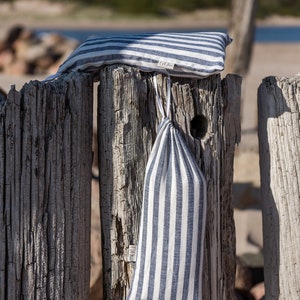 May include: A white and blue striped linen pillow and matching drawstring bag are hanging on a weathered wooden fence post. The pillow has a small white tag with the word "G&Ouml;Z" on it.